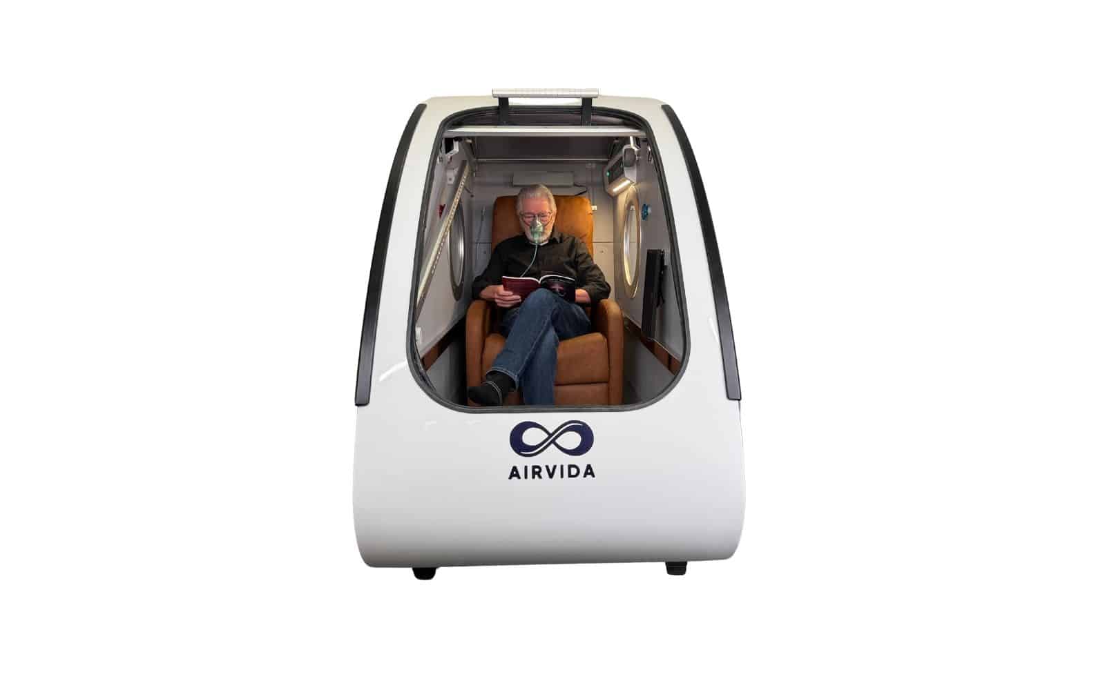 Patient seated comfortably inside a modern Airvida hyperbaric oxygen therapy chamber reading a book while wearing an oxygen mask.